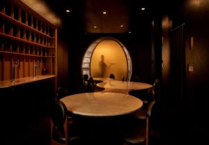 A cozy restaurant dining area with a large round table, wine glasses, and a server, surrounded by wood and glass elements, with soft and warm lighting