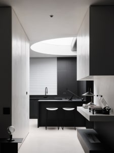 A dimly lit modern kitchen with a black counter and sink under a window, surrounded by three black and white chairs