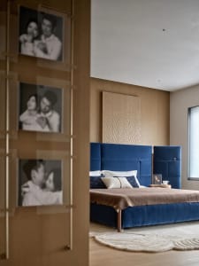 A modern bedroom with a blue bed, wooden headboard, and wooden floor, lit by natural light