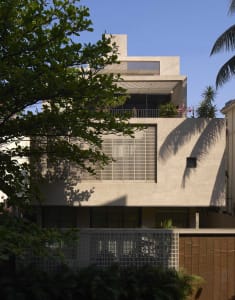 A modern house with a white exterior, large window, and balcony, surrounded by trees and greenery, with natural light streaming in through the window