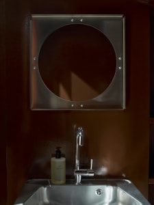 Small private bathroom with a metal sink and circular mirror, brown color scheme, and natural lighting