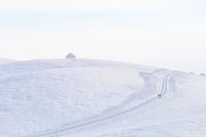 A snowy landscape with a road going up a hill, featuring two people walking and a car in the distance