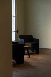 Black and white photograph of a minimalist living space with a wooden floor, a black chair, and a black bench, featuring a window in the background
