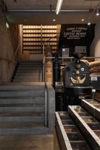 A modern coffee shop with a staircase leading to the second floor, surrounded by rustic coffee beans and a large coffee roaster