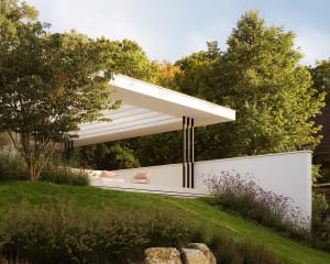 A modern house with a white exterior, situated on a hillside, surrounded by lush greenery and trees, with a large patio and a white roof