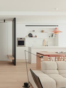 A modern kitchen and living space with a white oven, sink, and dining table, a couch and chair in the living room, and natural light from the windows