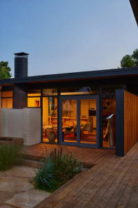 A modern house with a large open living room and kitchen, featuring a fireplace, couch, dining table, and refrigerator, with a black exterior and wooden deck, well-lit for a warm atmosphere
