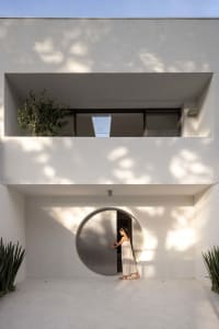 A woman stands in front of a modern white building with a circular window, surrounded by a calm and serene atmosphere