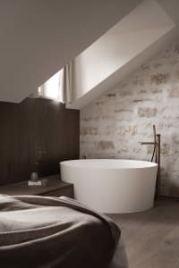 Small bathroom with a white bathtub, sink, and window, set against a stone wall and wooden floor in a dimly lit space