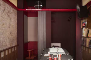 A small, dark room with a red wall, a black counter, and a few chairs, featuring cups and bottles on the counter