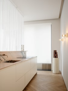 A modern kitchen with a white sink, cabinets, and marble countertop, set against a wooden floor, with natural light streaming in through the window
