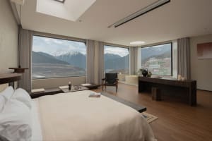 A bedroom with a large window offering a view of the mountains, featuring a bed, chair, desk, and potted plant, with a cozy atmosphere and warm natural light
