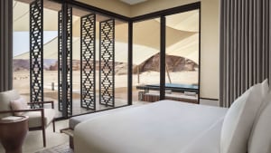 A modern hotel bedroom with a bed, chair, and large window, featuring wood and glass materials and natural light