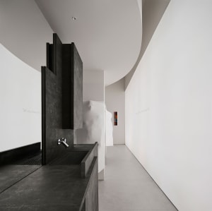 Bathroom with a sink on the left and a shower on the right, surrounded by white tiles and a dimly lit hallway in the background