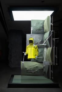 A mannequin in a yellow raincoat stands in front of a display case in a bright and inviting store or display area with a green backdrop
