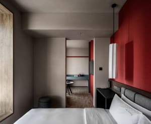 A warm and inviting hotel bedroom with a red-painted room, a black and white color scheme, and a bed positioned against a red wall, accompanied by a table and chair