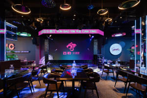 A well-lit restaurant interior with dining tables, chairs, and a large screen, surrounded by bottles and creating a comfortable atmosphere