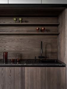 Modern kitchen with wooden cabinets, a black sink, and a wooden countertop, featuring a shelf with glasses and a dimly lit atmosphere