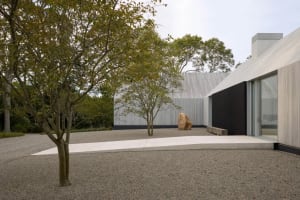 A modern house with a stone exterior and a large rock in the front yard, set in a calm and serene outdoor setting with a patio area visible