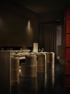 Dimly lit dining room with a wooden floor, a dining table, chairs, wine glasses, and a vase, creating a cozy atmosphere