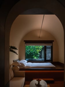Cozy bedroom with a bed under a window, a lamp on the left side, and a brick wall adding a rustic touch