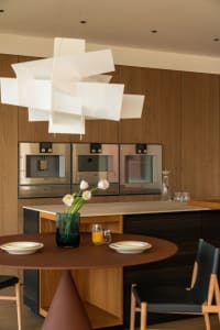 A modern kitchen with wooden cabinets, a dining table, and a chair, set with plates, cups, and a vase of flowers, under natural light