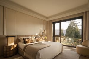 A well-lit bedroom with a bed near the window, a chair nearby, and a beige color scheme creating a warm atmosphere