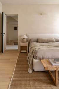 A modern bedroom with a wooden floor, a white wall, a bed with a white blanket and brown headboard, a table, and a chair, lit by natural sunlight