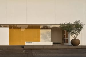 Photograph of a modern building with a white exterior, a yellow door, and a large glass window allowing natural light to enter the space