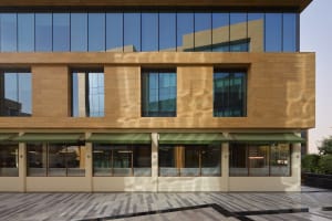 Photograph of a modern building with a tan brick facade, a large window, and a green awning, bathed in natural sunlight