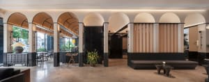 Well-lit modern lobby with a black and white color scheme, featuring a couch, dining table, bench, and potted plants, with a large archway leading to another room