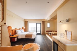 A modern bedroom with a bed, chair, and dining table, featuring wooden floors and natural light from a window