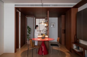 A modern kitchen with a woman standing at a red dining table, surrounded by chairs, a bowl, and a potted plant, with a sink and refrigerator, featuring wood and glass materials and natural lighting