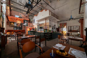 Cozy restaurant interior with tables, chairs, and a bar, featuring potted plants, vases, and a bowl of fruit, with a mix of natural and artificial lighting
