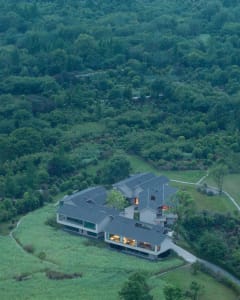 A modern house with a black roof, surrounded by a lush green field, blending indoor and outdoor living spaces with a patio and balcony, bathed in natural sunlight