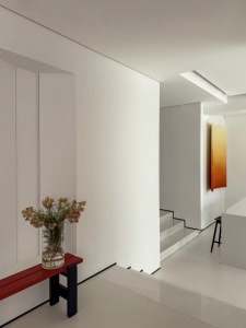 A well-lit living room or dining area with a red bench, a vase with flowers, and a staircase in the background, set against a predominantly white background