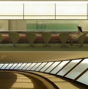 A modern lobby with a curved wall and a row of wooden chairs in a semi-circle, with a person sitting in one of them, and a large window providing natural light