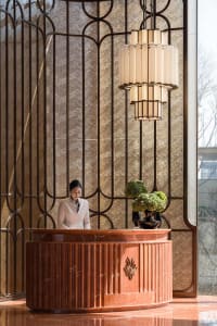 A woman stands behind a marble counter in a dimly lit hotel lobby with a chandelier and elegant atmosphere