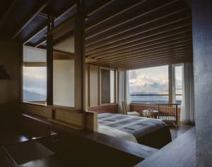 A dimly lit bedroom with a wooden floor, a wooden headboard, and a bed positioned near the window, accompanied by a chair