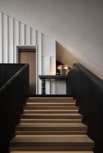 A black and white photograph of a wooden staircase with a black railing, lit by a light source creating a contrast between the dark staircase and the lit area