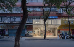 A modern restaurant with a glass and concrete facade, a large window, and a door, set against a backdrop of a tree and soft, diffused lighting