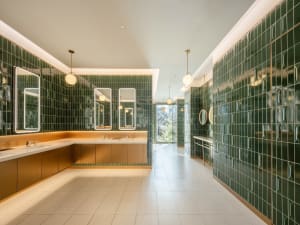 A bathroom with a white tile floor and green tile walls, featuring a sink, mirror, and window, with soft warm lighting