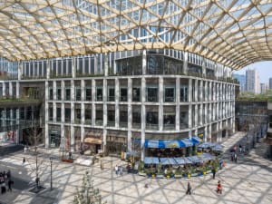 A large open space with a glass ceiling, natural light, and people walking around, with umbrellas placed throughout, indicating an outdoor event or gathering