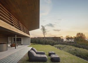 Modern house with a large deck and patio, featuring black lounge chairs and a grassy area, with natural light pouring in through a large window