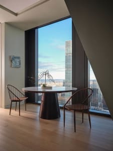 Cozy modern living space with a dining table, chairs, and a potted plant, surrounded by a large window with a city view, and well-lit with natural light