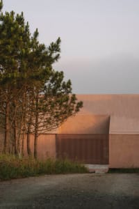 A modern building with a pink exterior, surrounded by trees, featuring a large door and window, with a concrete floor and a bench in a serene outdoor setting