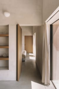 A cozy bedroom with a wooden floor, a white wall, a bed, a bookshelf, and a vase on the bookshelf, with natural light entering through an open door