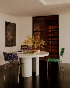 A well-lit living room with a white table, chairs, a vase with flowers, a dining table, and a potted plant, featuring a combination of wood and glass materials