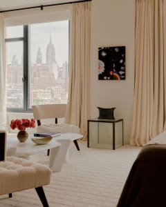 A modern living room with a large window overlooking the city, featuring a couch, chair, and dining table, with a vase and potted plant adding greenery to the space, lit by natural light