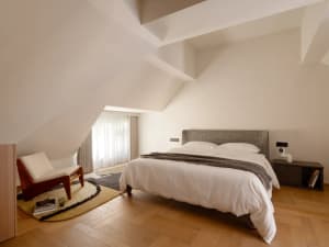 A simple bedroom with a wooden floor, a bed, a chair, and a desk, with a focus on comfort and functionality